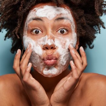 People, wellness, hygiene and skin problems concept. Lovely Afro American lady keeps lips folded, touches cheeks, has white foam on faces, washes with beauty gel, feels refreshed, has wide opened eyes
